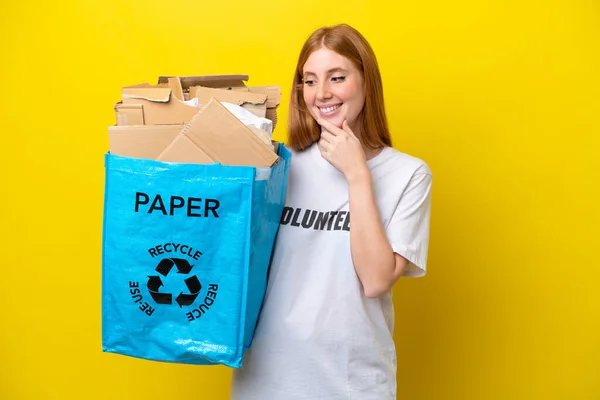 リサイクルバッグを持っている若い赤毛の女性側を見て黄色の背景に隔離されたリサイクルすると笑顔 — ストック写真