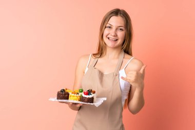 Ukraynalı aşçı pembe arka planda izole edilmiş bir kek tutarak telefon jesti yapıyor.