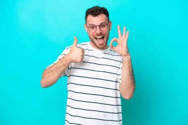 Mavi arka planda izole edilmiş genç Brezilyalı adam işaret ve baş parmak hareketi gösteriyor.