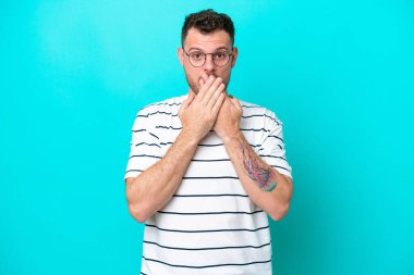 Mavi arka planda izole edilmiş genç Brezilyalı adam elleriyle ağzını kapatıyor.