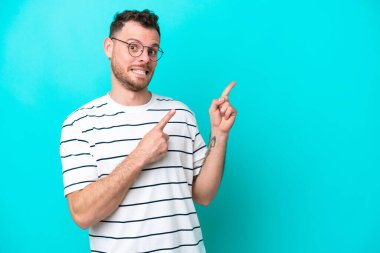 Mavi arka planda izole edilmiş genç Brezilyalı adam korkmuş ve kenarı işaret ediyor.