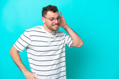 Genç Brezilyalı bir adam mavi arka planda izole edilmiş bir şekilde elini kulağına koyarak bir şeyler dinliyor.
