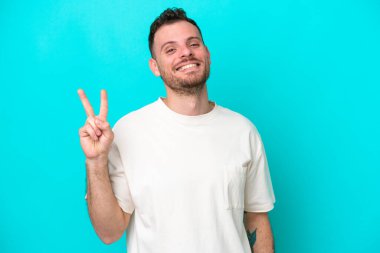 Mavi arka planda izole edilmiş genç Brezilyalı adam gülümsüyor ve zafer işareti gösteriyor