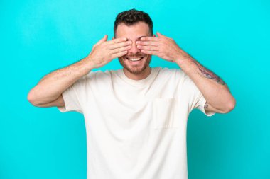 Genç Brezilyalı bir adam mavi arka planda izole edilmiş. Gözlerini elleriyle kapatıyor ve gülümsüyor.