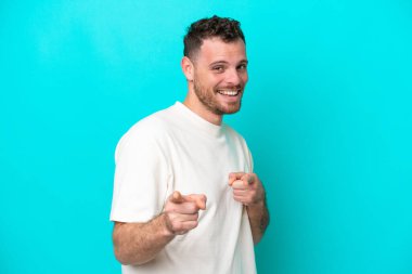 Mavi arka planda izole edilmiş genç Brezilyalı adam ön tarafı işaret ediyor ve gülümsüyor.