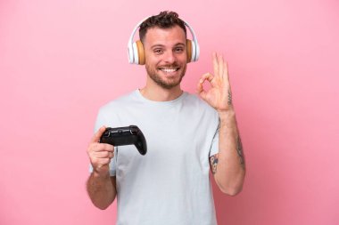 Pembe arka planda izole edilmiş bilgisayar oyunu kontrolörüyle oynayan genç Brezilyalı adam parmaklarıyla tamam işareti gösteriyor