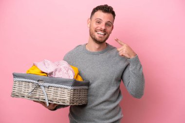 Genç Brezilyalı bir adam pembe arka planda izole edilmiş bir çamaşır sepeti tutuyor ve baş parmak işareti yapıyor.