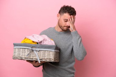 Genç Brezilyalı bir adam pembe arka planda izole edilmiş, yorgun ve hasta bir ifadeyle bir çamaşır sepeti tutuyor.