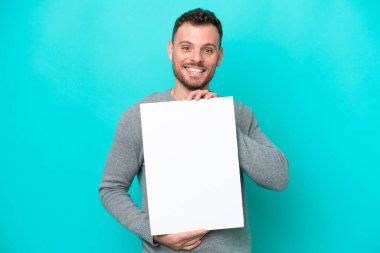 Elinde boş bir levha tutan genç Brezilyalı adam mavi arka planda izole edilmiş mutlu bir ifadeyle boş bir pankart tutuyor.