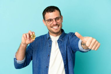 Elinde Bitcoin tutan Brezilyalı bir adam izole edilmiş mavi arka planda baş parmak işareti yapıyor.