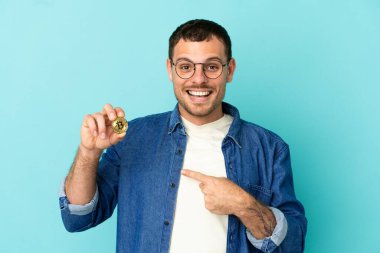 Brezilya 'lı bir adam Bitcoin' i izole edilmiş mavi arkaplanda tutuyor ve şaşırmış yüz ifadesi var.