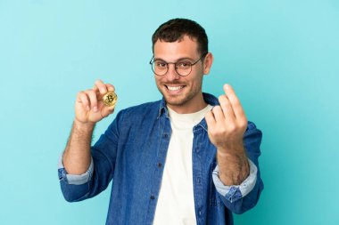 Elinde Bitcoin olan Brezilyalı bir adam izole edilmiş mavi arka planda para hareketi yapıyor.