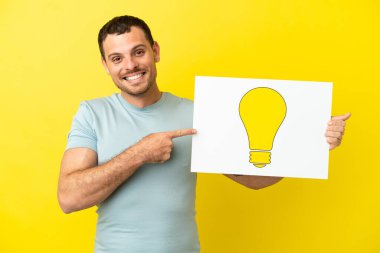 İzole edilmiş mor arka plan üzerinde Brezilyalı bir adam elinde ampul simgesi olan bir levha tutuyor ve ona doğrultuyor.