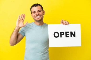 Mor arkaplan üzerinde Brezilyalı bir adam. Elinde açık yazılı bir pankart var ve selam veriyor.