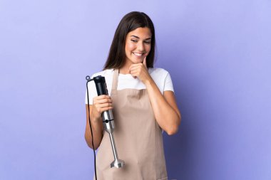 Mor arka planda izole edilmiş el blenderi kullanan Brezilyalı kadın yana bakıyor ve gülümsüyor.