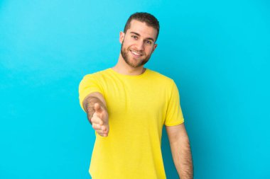 Mavi arka planda izole edilmiş yakışıklı beyaz adam iyi bir anlaşma için el sıkışıyor.