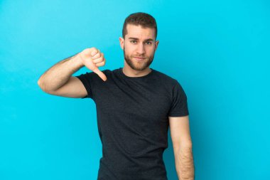Mavi arka planda izole edilmiş genç, yakışıklı beyaz adam, olumsuz bir ifadeyle baş parmağını aşağı gösteriyor.