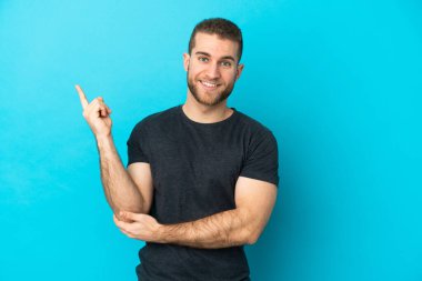 Mavi arka planda izole edilmiş yakışıklı beyaz adam mutlu ve dik duruyor.