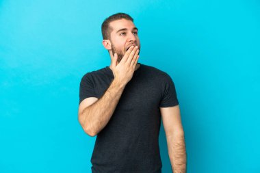 Mavi arka planda izole edilmiş yakışıklı beyaz adam esniyor ve ağzını eliyle açık tutuyor.