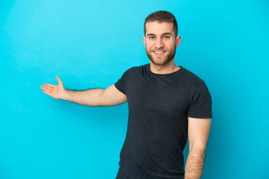 Mavi arka planda izole edilmiş yakışıklı beyaz adam, davet edilmek için ellerini yana uzatıyor.