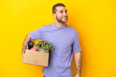 Kafkasyalı genç, sarı arka planda izole edilmiş eşyalarla dolu bir kutuyu kaldırırken yukarı bakarken bir fikir düşünüyor.