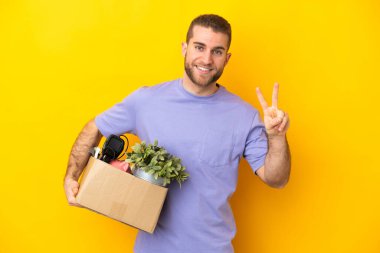Genç Kafkasyalı, sarı arka planda izole edilmiş şeylerle dolu bir kutuyu alırken gülümsüyor ve zafer işareti gösteriyor.