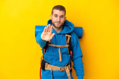 Sarı arka planda izole edilmiş büyük sırt çantalı beyaz bir adam dur hareketi yapıyor.