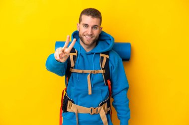 Sarı arka planda izole edilmiş büyük bir sırt çantası olan beyaz dağcı gülümsüyor ve zafer işareti gösteriyor.