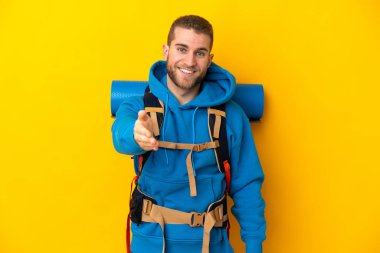 Sarı arka planda izole edilmiş büyük sırt çantalı beyaz bir dağcı iyi bir anlaşma için el sıkışıyor.