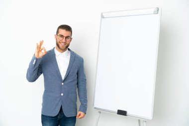 Beyaz arka planda izole edilmiş beyaz panoda sunum yapan genç iş kadını parmaklarıyla tamam işareti gösteriyor.