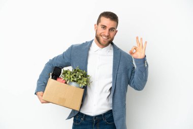 Beyaz arka planda izole edilmiş eşyalarla dolu bir kutuyu alırken, beyaz tenli genç hareket ediyor. Parmaklarıyla tamam işareti gösteriyor.