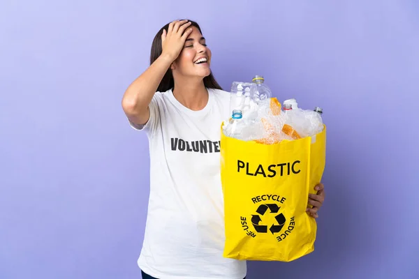 若いブラジル人女性が袋を持っていますプラスチックボトルでいっぱい紫色の背景に隔離されたリサイクル多くの笑顔 — ストック写真