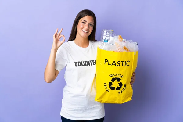 若いブラジル人女性を保持バッグフルプラスチックボトルリサイクルに隔離された紫色の背景に示すOkサインで指 — ストック写真