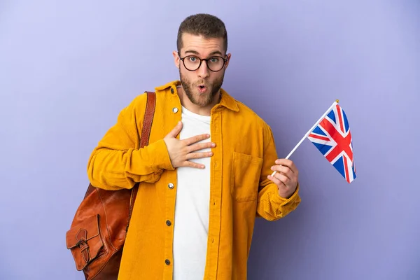 年轻的高加索男子举着一面英国国旗 背景是紫色的 眼睛看着右边 感到惊讶和震惊 — 图库照片