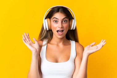 Sarı arka planda izole edilmiş genç beyaz kadın şaşırdı ve müzik dinliyor.