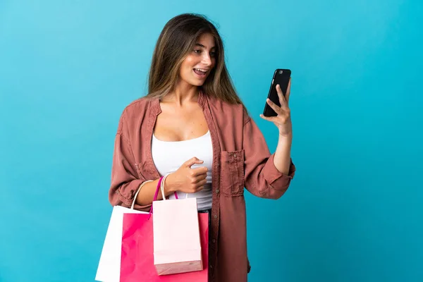 青の背景に隔離された若い白人女性ショッピングバッグを保持し 友人に彼女の携帯電話でメッセージを書く — ストック写真