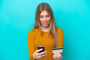 Genç beyaz kadın mavi arka planda izole edilmiş cep telefonuyla kredi kartıyla alışveriş yapıyor.