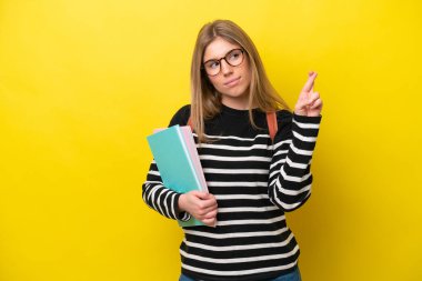 Sarı arka planda izole edilmiş genç bir öğrenci kadın parmaklarını çapraz yapıp en iyisini diliyor.