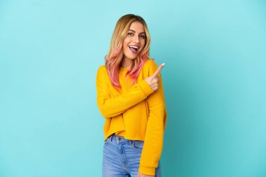 İzole edilmiş mavi arka planı olan genç bir kadın şaşırmış ve işaret ediyor.