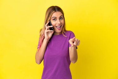 İzole edilmiş sarı arka plan üzerinde cep telefonu kullanan genç bir kadın şaşırmış ve ön tarafı işaret ediyor.