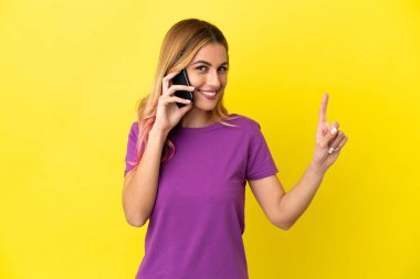 İzole edilmiş sarı arka plan üzerinde cep telefonu kullanan genç bir kadın en iyinin işareti olarak parmağını kaldırıyor.