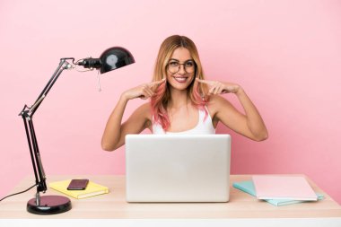 İşyerindeki pembe arka planda dizüstü bilgisayarı olan genç bir bayan baş parmak hareketi yapıyor.