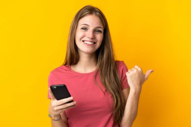 Sarı arka planda izole edilmiş cep telefonu kullanan genç bir kadın ürünü sunmak için yan tarafı işaret ediyor.