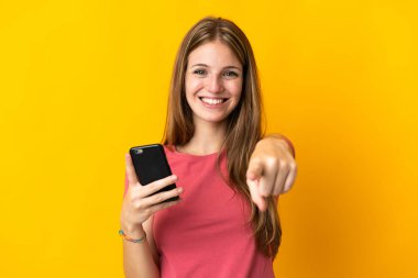 Sarı arka planda izole edilmiş cep telefonu kullanan genç bir kadın kendine güvenen bir ifadeyle seni işaret ediyor.
