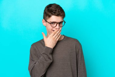 Genç Rus adam mavi arka planda izole edilmiş mutlu ve gülümseyen ağızlarını eliyle kapatıyor.