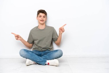 Genç Rus adam beyaz arka planda izole bir şekilde yerde oturuyor. Parmak uçlarını işaret ediyor ve mutlu.