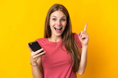 Sarı arka planda izole edilmiş cep telefonu kullanan genç bir kadın harika bir fikre işaret ediyor.