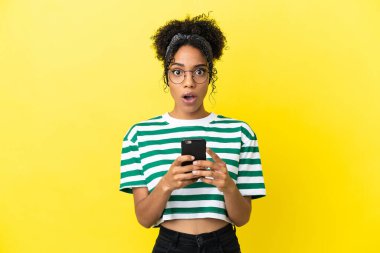 Genç Afrikalı Amerikalı kadın sarı arka planda izole edilmiş kameraya bakarken şaşırmış bir ifadeyle cep telefonunu kullanıyor.