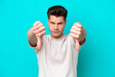 Genç, yakışıklı, beyaz bir adam, mavi bir bakkalda izole edilmiş, baş parmağını iki eliyle aşağı gösteriyor.