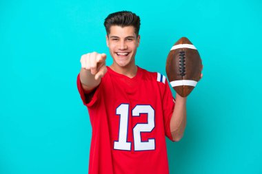 Rugby oynayan beyaz bir adam mavi arka planda izole edilmiş, şaşırmış ve ön tarafı işaret ediyor.